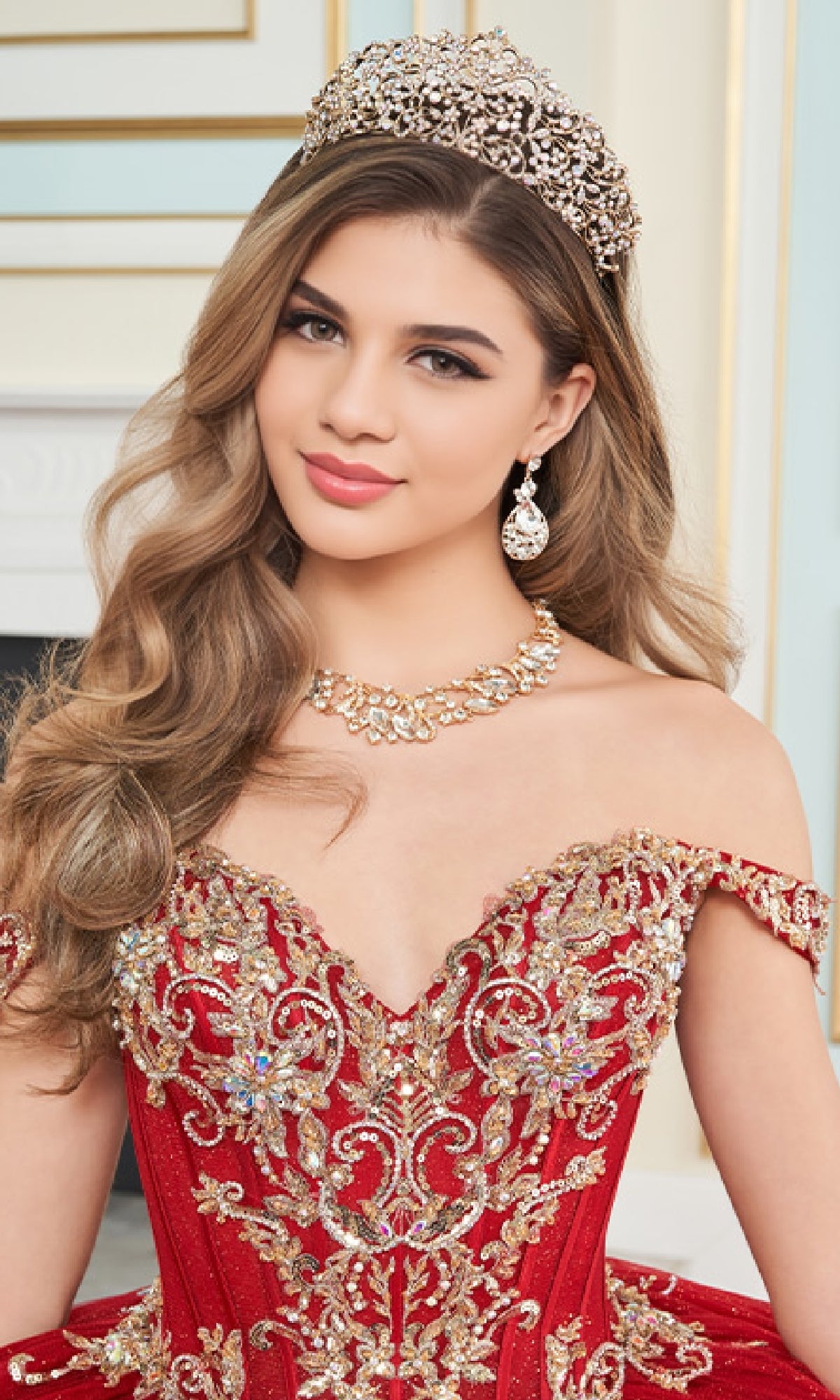 A young woman poses indoors in the Quinceanera Dress PR30171 by Princesa—an off-the-shoulder red and gold embroidered ball gown with glitter tulle, paired with a jeweled tiara and ornate gold necklace and earrings.